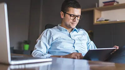 Guide to Data Analytics: The Trend That's Reshaping the Insurance Industry Man in business attire sitting behind a desk in an office, he is looking down at a tablet in his hands.